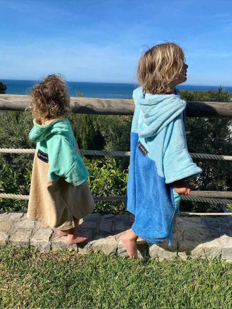 Doi copii imbracati in poncho plaja copii - unul de culoare crem si turquoise, iar altul de culoare albastru