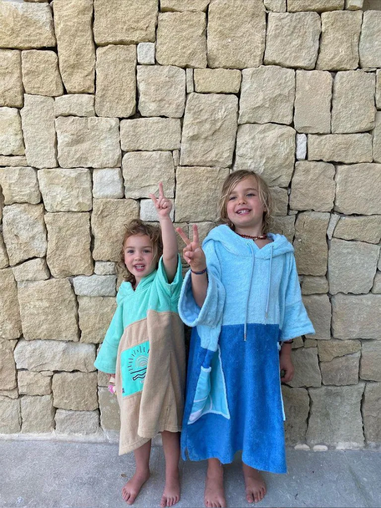 Doi copii imbracati in poncho plaja copii - unul de culoare crem si turquoise, iar altul de culoare albastru